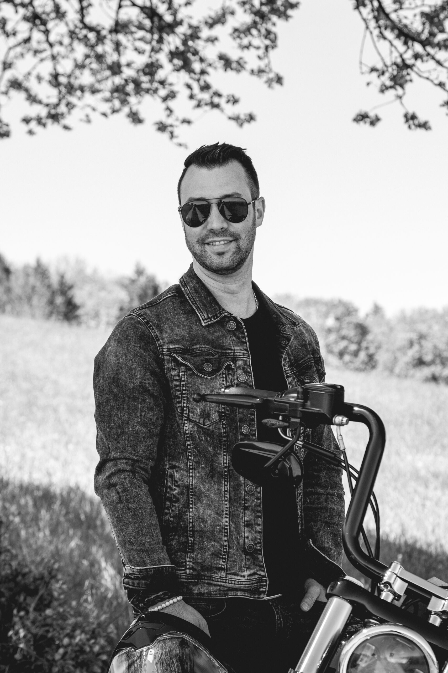 Herr steht neben seinen Motorrad und hat Sonnenbrille auf.
Bild in Schwarz/Weiß